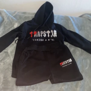 Svart hoodie från Trapstar - Säljer en svart hoodie från Trapstar med tryck i vitt och rött framtill. Hoodien har magficka och justerbar huva. Perfekt för en avslappnad streetwear-look. tracksuit 