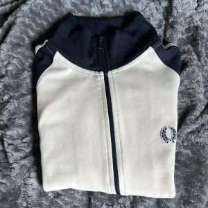 Vit och marinblå track jacket från Fred Perry - Säljer en stilren vit och marinblå track jacket från Fred Perry med dragkedja och broderad logga på bröstet. Jackan har ribbade muddar och klassisk sportig look. Perfekt för dig som gillar retrostil och streetwear.