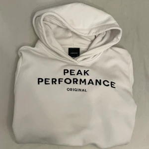 Vit hoodie från Peak Performance - Säljer en vit hoodie från Peak Performance med svart broderad logga på bröstet. Tröjan har huva och en stor magficka framtill. Lite defekter, kan försvinna med Vanish. Perfekt för en avslappnad och sportig stil. 170 men Passar XS/S