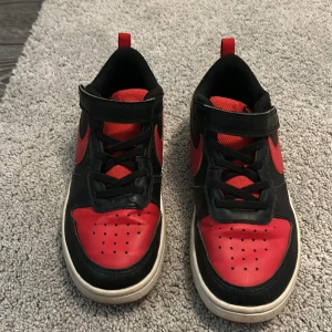 Nike sneakers i svart och rött - Säljer ett par klassiska Nike sneakers i svart och rött med vit sula. Skorna har snörning och kardborreband för extra bra passform. Perfekta för dig som gillar sportig stil och vill sticka ut med färg. Ikonisk Nike-logga på sidan.