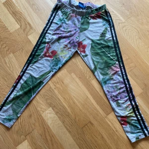 Leggings från Adidas  - Säljer ett par grå leggings från Adidas med färgglatt blommönster och svarta ränder längs sidorna. Byxorna har elastisk midja och Adidas-logga på låret. Perfekta för dig som gillar sportig stil med twist.