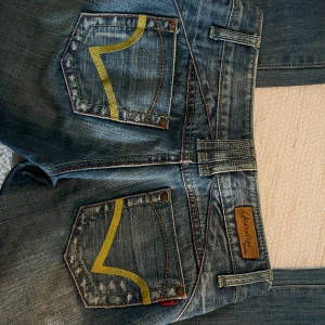 Lågmidjade Blåa jeans från Miss Sixty  - Säljer ett par blå jeansbyxor från Miss Sixty med snygga slitningar och kontrastsömmar i rött. Bakfickorna har en gul broderad detalj som sticker ut. 