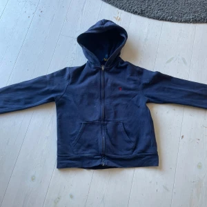 Mörkblå hoodie från Polo Ralph Lauren - Säljer en mörkblå hoodie från Polo Ralph Lauren med dragkedja och huva. Tröjan har två fickor framtill och en liten röd logga broderad på bröstet. Perfekt för en avslappnad stil.