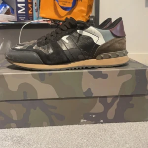Valentino Rockrunner sneakers med camouflage - Säljer ett par Valentino Rockrunner sneakers med svart, grått och grönt camouflage-mönster. Skorna har svarta snören, rund tå och detaljer i mocka och skinn. Sulan är beige med nitar på hälen och baksidan har en vinröd detalj.  Skick 7/10 lite slitage där bak i skon men inget som syns när den är på 🙌🙌
