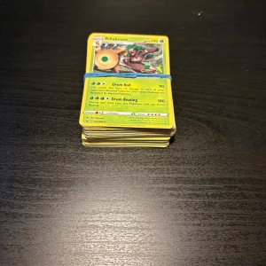 Pokémonkort bunt med Rillaboom på toppen - Säljer en bunt Pokémonkort där Rillaboom är det översta kortet. Korten är samlade i en hög och hålls ihop med ett blått gummiband. Perfekt för dig som vill utöka din samling med olika Pokémonkort.
