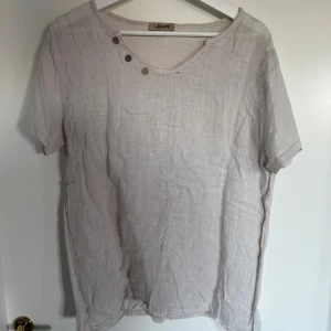 Beige t-shirt med knappar vid halsen - Säljer en beige bohemisk t-shirt med unik knappdetalj. T-shirten har en avslappnad passform. Perfekt för en stilren och enkel look. 