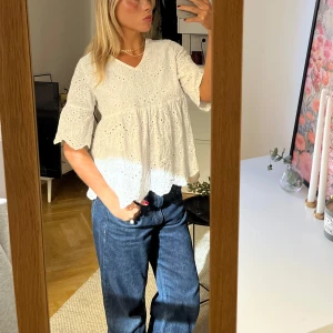 Vit blus med broderade detaljer - Säljer en söt vit blus med broderade hålmönster och volangkant. Blusen har korta ärmar och en lös passform, perfekt för vår och sommar. Den har en v-ringad halsringning.