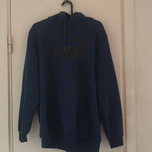 Blå hoodie från Puma - Mörkblå hoodie från Puma med klassisk logga framtill. Tröjan har huva med dragsko och en stor magficka. Perfekt för en avslappnad stil.