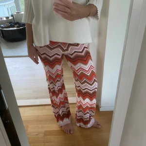 Mönstrade stickade byxor i- missoni liknande  - Säljer ett par stickade byxor med färgglatt zickzackmönster i rött, rosa, brunt och beige med glitter. Byxorna har en loose passform och är perfekta för dig som gillar en unik och avslappnad stil. 😍Märke An’ge- Stolek One size- Nypris ca 800