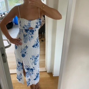 Vit och blå blommig långklänning med slits - Säljer en vit långklänning med blått blommönster och smala axelband. Klänningen har en liten cut-out framtill och en slits framtill för extra detalj. Perfekt för sommarens alla tillfällen! Endast använd på midsommar förra året😍