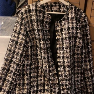 Svartvit rutig kappa - Säljer en snygg svartvit kappa med rutigt mönster. Jackan har lång ärm och är öppen framtill utan synliga knappar eller dragkedja. Perfekt för dig som vill ha en stilren och trendig look.