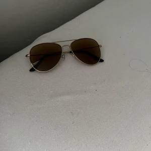 Guldiga pilotbågar solglasögon - Snygga solglasögon med klassisk pilotform och tunna guldiga metallbågar. Glasen är bruna och bågarna har svarta detaljer vid ändarna. Perfekta för en stilren look i solen.