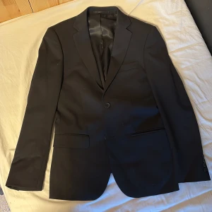 Svart kostym från Dressmann Slim Fit - Säljer en svart tvådelad kostym från Dressmann i slim fit-modell. Kavajen har klassisk design med två fickor och en bröstficka, samt snyggt röd kantband på insidan. Byxorna är raka och matchar kavajen perfekt.