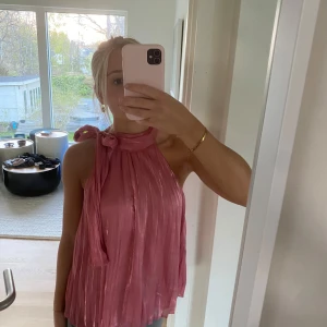 Rosa plisserad halterneck blus med rosett - Säljer en glansig rosa plisserad halterneck blus med stor rosett vid halsen.  Blusen är ärmlös och har ett luftigt, flowy fall. Perfekt för dig som vill sticka ut med en feminin och trendig topp! Från Mango och nypris 499kr🥰😍Endast använd en gång! 