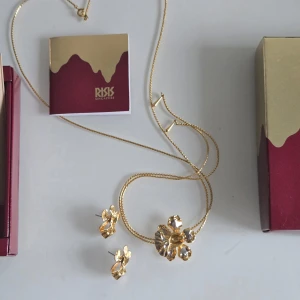 halsband och örhängen  RISIS - Säljer ett vackert set från RISIS med halsband och örhängen i guldfärg, båda med detaljer i form av blommor. Perfekt för dig som vill ha ett unikt och elegant smycke med naturinspirerad design. Kedjan är tunn och smidig, och örhängena är i matchande stil.