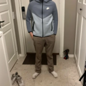 Ljusblå Nike tech fleece - Säljer en ljusblå och grå hoodiejacka från Nike med dragkedja och huva. Jackan har svart Nike-logga på bröstet och svarta detaljer vid dragkedjan. Perfekt för en sportig och avslappnad stil. Det finns lite små hål men inget man bryr sig om