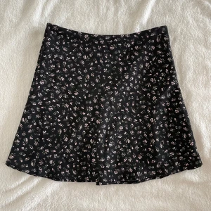 Svart blommig kjol från h&m - Supersöt svart kjol från h&m med små blommor i vitt och rosa. Kjolen har en lätt utställd form och är kort i modellen. Perfekt att styla med en enkel topp eller hoodie. Midja: 28cm, Längd: 35cm