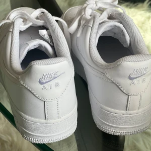 Vita Nike Air Force 1 sneakers - Säljer ett par klassiska vita Nike Air Force 1 sneakers. Skorna har en stilren design med helvit ovandel, snörning och Nike-logga bak på hälen. Perfekta för dig som gillar en clean och tidlös look.