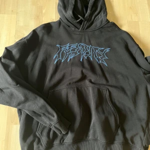 Svart hoodie med blått tryck - Säljer en svart hoodie med ett blått grafiskt tryck på bröstet. Tröjan har huva och en stor magficka framtill. Perfekt för dig som gillar streetwear och vill ha en bekväm och snygg tröja.