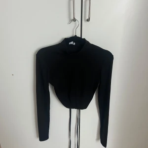 Svart croppad långärmad topp med knyt - Säljer en svart croppad topp med lång ärm och hög krage. Toppen har knytband i midjan som ger en snygg detalj och markerar midjan. Perfekt att styla till både jeans och kjol.
