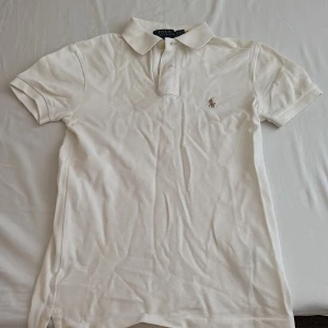 Vit piké från Polo Ralph Lauren - Klassisk vit pikétröja från Polo Ralph Lauren med broderad logga på bröstet. Slim fit-modell med korta ärmar och krage med knappar. Perfekt för en stilren och tidlös look.