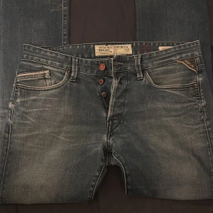 Blå Replay Waitom jeans - Säljer ett par blå Replay Waitom jeans med klassisk femficksdesign och snygga slitningar. Jeansen har knappar i gylfen och en diskret Replay-lapp vid fickan. Passar dig som gillar en tidlös jeanslook. Har ett litet hål mellan benen.