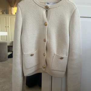 Beige stickad kofta från Zara - Säljer en beige stickad kofta från Zara med guldknappar framtill och två fickor. Koftan har lång ärm och en klassisk, rak passform. Perfekt att slänga över en topp för en stilren look. Har andvänst endast 1-2 gånger eftersom att den är lite för liten för mig, storleken är S och är i grymt skick!