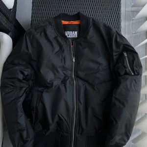 Svart bomberjacka från Urban Classics - Säljer en svart bomberjacka från Urban Classics med orange foder. Jackan har dragkedja framtill, fickor på sidorna och en extra ficka med dragkedja på ärmen. Klassisk modell med ribbade muddar vid ärmslut och midja. Storlek XL.