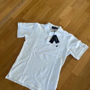 Ralph lauren pike - Klassisk vit pikétröja från Polo Ralph Lauren med broderad logga på bröstet. Tröjan har krage, knappar vid halsen och är kortärmad. Perfekt för en stilren och tidlös look. Helt ny o har etiketten på. Storlek S. 