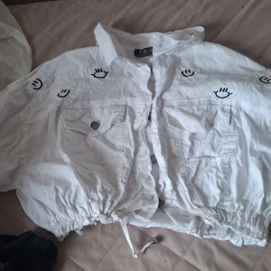 Vit cropped jacka med broderade smileys - Säljer en vit cropped jacka från Cobra med broderade svarta smileys i hjärtan över hela jackan. Jackan har dragsko i midjan, stora bröstfickor med knapp och långa ärmar. Perfekt för dig som vill sticka ut med en unik stil.