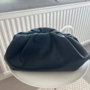 Svart clutchväska  - Säljer en svart clutchväska med mjuk, puffig design och rynkad öppning. Väskan är tillverkad i skinnimitation och har ett stilrent, minimalistiskt utseende. Perfekt för att bära det viktigaste när du vill ha en snygg accessoar. Från Carin Wester. 