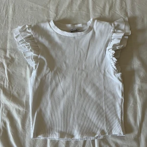Vit topp med volang ärmar  - Säljer en söt vit ribbad topp från Zara med volangdetaljer på ärmarna. Denna är helt oanvänd då den tyvärr är lite för kort för mig. Det är storlek M men passar s/xs beroende på hur man vill ha passformen.