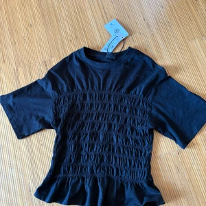 Svart smockad topp med volangkant - Säljer en svart topp med smockad midja och volangkant nertill. Toppen har rund halsringning och korta ärmar. Perfekt för dig som vill ha en trendig och bekväm look. prislappen är fortfande kvar och jag väljer att sälja den för att jag råka beställa två. skriv för fler frågor💕