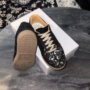 Svarta sneakers med färgstänk från Maison Margiela - Unika svarta sneakers från Maison Margiela med vita snören och färgstänk i blått, vitt, gult och orange. Skorna har rund tå och platt sula, samt en klassisk siluett. Perfekta för dig som vill sticka ut med din stil.