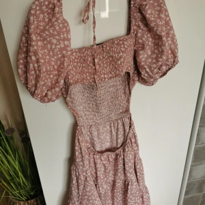 Rosa blommig klänning med puffärmar - Supersöt rosa klänning med små vita blommor och puffiga kortärmar. Smockad upptill och i midjan för en bekväm passform. Klänningen har en romantisk känsla och knytband i nacken.