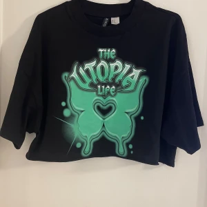 Svart croppad t-shirt med grön tryck - Cool svart croppad t-shirt från H&M Divided  med stort grönt tryck framtill där det står 'The Utopia Life'. Oversized passform och korta ärmar, perfekt till streetwear eller en avslappnad look.