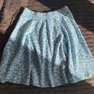 Blommig blå kjol med resårmidja - Säljer en söt blå kjol med små vita blommor. Kjolen har en lätt utställd form och är perfekt för varma dagar. Tunt och mjukt tyg som ger en skön känsla.