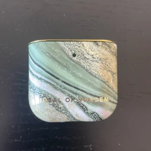 Grön och guldig AirPods-fodral från iDeal of Sweden - Snyggt AirPods-fodral i marmorerat mönster med gröna, vita och guldiga toner. Fodralet har en lyxig känsla med gulddetaljer och passar perfekt för att skydda dina hörlurar med stil.