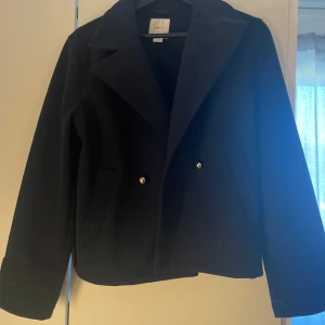 Svart kappa med guldknappar - Stilren svart kappa med klassisk krage och två guldiga knappar framtill. Jackan har lång ärm och två fickor på sidorna. 