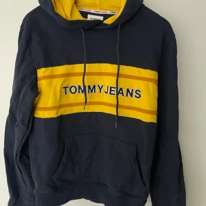 Mörkblå hoodie från Tommy Jeans med gul detalj - Säljer en mörkblå hoodie från Tommy Jeans med gul huva och breda gula ränder över bröstet. Stor logga framtill och klassisk känguruficka. Perfekt för en avslappnad och sportig stil. XS men kan användas som S 