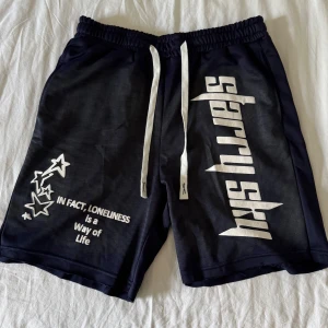 Svarta shorts med tryck och snörning - Riktigt feta och snygga shorts med vitt tryck på ena benet och texten 'IN FACT, LONELINESS is a Way of Life'. Har vita dragsnören i midjan och elastisk resår. Perfekta för en avslappnad stil.