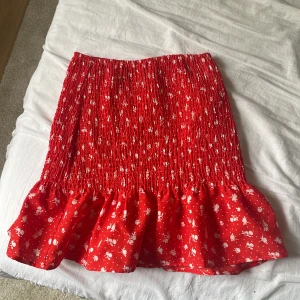 Röd blommig kjol med volang - Supersöt röd kjol från H&M med små vita blommor och volang nertill. Kjolen är kort och har en smockad midja. Den går även att använda som en topp. Aldrig använd och i storlek XXS.