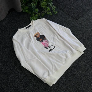 Vit sweatshirt från Polo Ralph Lauren - Vit sweatshirt från Polo Ralph Lauren med ett stort tryck av en björn på framsidan. Tröjan har rund halsringning och långa ärmar. Tillverkad i 100% bomull och har RLX-tryck under motivet.