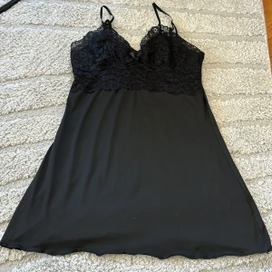 Svart spetsklänning/nattlinne med tunna axelband - Säljer en svart klänning med spets upptill och tunna justerbara axelband. Klänningen har en liten rosett framtill och är ärmlös med en mjuk, slät kjoldel. Perfekt för dig som gillar en feminin och elegant stil.