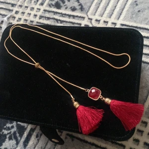Guldfärgat halsband med röda tofsar och sten - Säljer ett halsband med tunn guldfärgad kedja. 