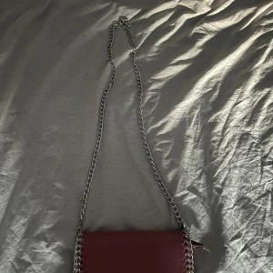 Rosa axelväska med kedja från Tiamo - Säljer en snygg rosa axelväska från Tiamo med silverfärgad kedjerem och dekorativa sömmar längs kanterna. Väskan har dragkedja och är perfekt för att bära det viktigaste. Stilren och trendig accessoar för dig som gillar färg.