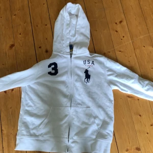 Vit hoodie från Polo Ralph Lauren med USA-tryck - Vit hoodie från Polo Ralph Lauren med dragkedja, fickor framtill och tryck med USA och siffra 3 på bröstet. Stor broderad logga och amerikansk flagga på ärmen. Perfekt för en sportig look.