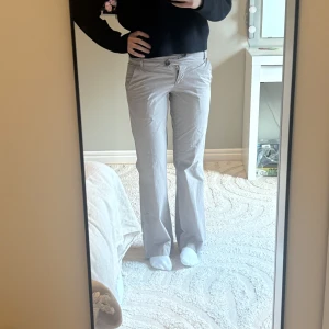 Lågmidjade beiga bootcut byxor med knappar - Snygga beiga byxor med bootcut-passform och två knappar i midjan. Byxorna har en låg midja. Byxorna har fickor fram och bak samt en klassisk look som passar till många stilar. Perfekta för dig som gillar en lite vidare modell nertill.