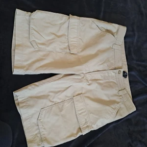 Beiga cargoshorts från Everest - Säljer ett par beiga cargoshorts från Everest med stora fickor på sidorna och en bakficka. Klassisk passform och tillverkade i bomull, perfekta för sommardagar. Stängs med knapp och dragkedja.