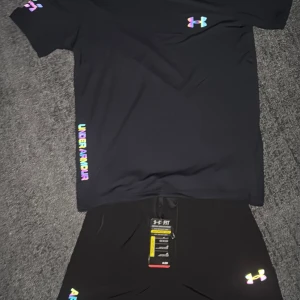 Svart t-shirt från Under Armour med regnbågsdetaljer - Svart t-shirt från Under Armour med färgglada loggor och textdetaljer i regnbågsfärger på bröstet, ärmen och sidan. Perfekt för träning eller sportiga outfits. Materialet känns lätt och andas bra.
